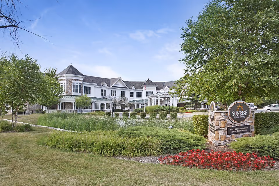 Exterior view of a senior living facility with a large white building featuring multiple windows and a covered entrance. The foreground includes well-maintained landscaping with green grass, bushes, trees, and a flower bed with red flowers. A stone sign on the right reads 'Sunrise Senior Living' with additional text below.