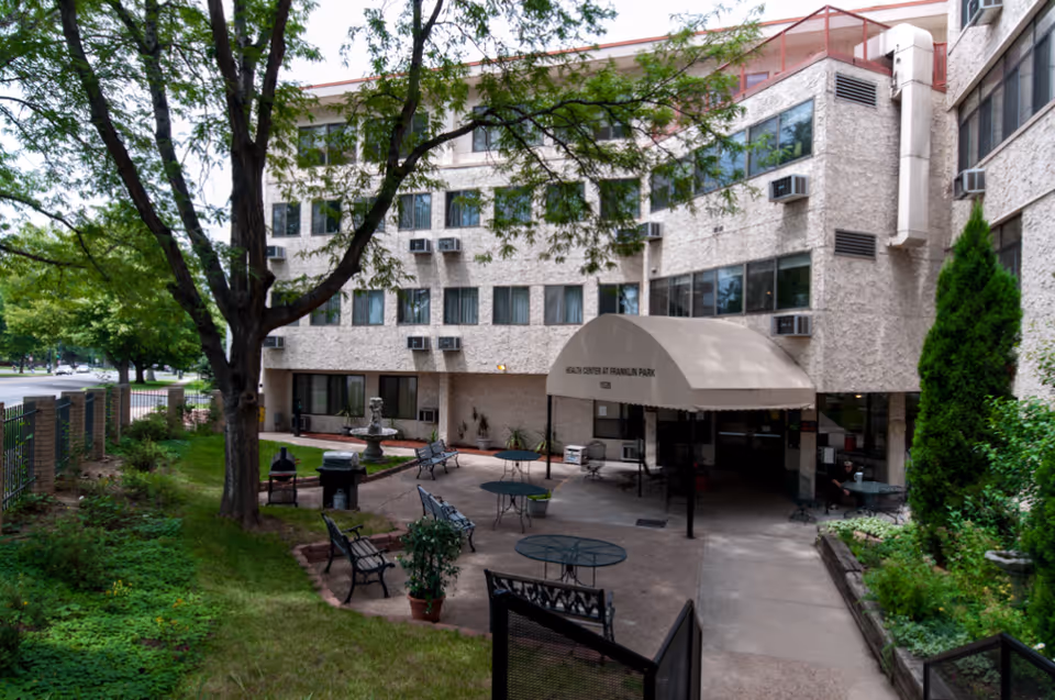 Outdoor courtyard area of a multi-story building with a beige textured exterior. The courtyard features several metal tables and chairs, benches, potted plants, a small fountain, and a canopy entrance labeled 'Health Center at Franklin Park'. Trees and greenery surround the area, and there is a sidewalk leading to the entrance.