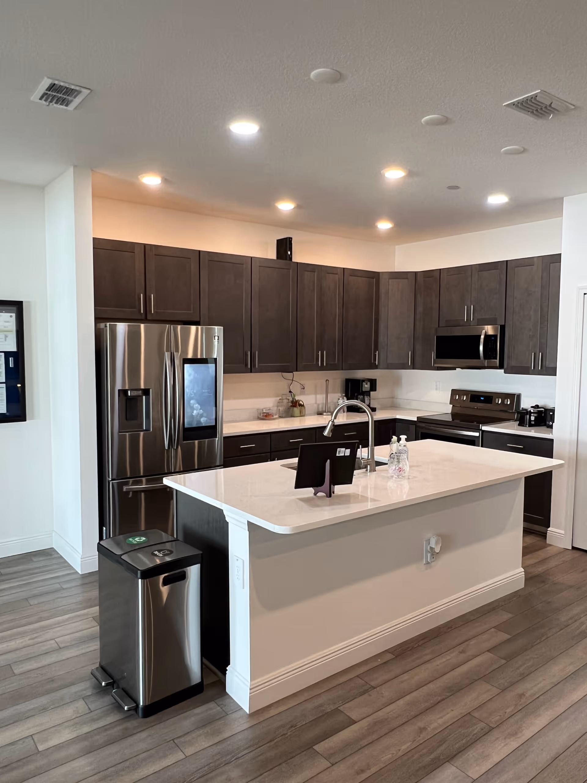 Modern kitchen area with dark wood cabinets, stainless steel refrigerator, stove, microwave, and a white island countertop with a sink and soap dispensers. There is a trash bin next to the island and wooden flooring throughout.