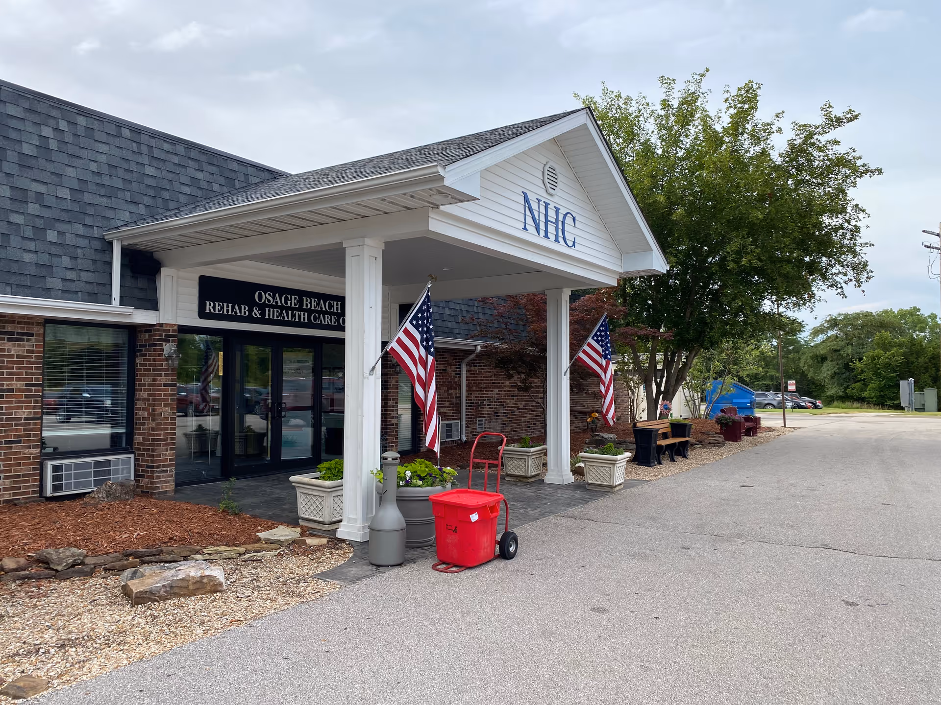 Front entrance of Osage Beach Rehab & Health Care Center showing a covered entry with columns, American flags, planters, and a red utility cart.