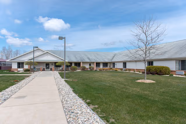 Exterior view of a single-story assisted living facility building with a long sidewalk leading to the entrance. The building has a gray roof, white siding, and several windows. There is a grassy lawn with a few bushes and a leafless tree on the right side under a partly cloudy sky.