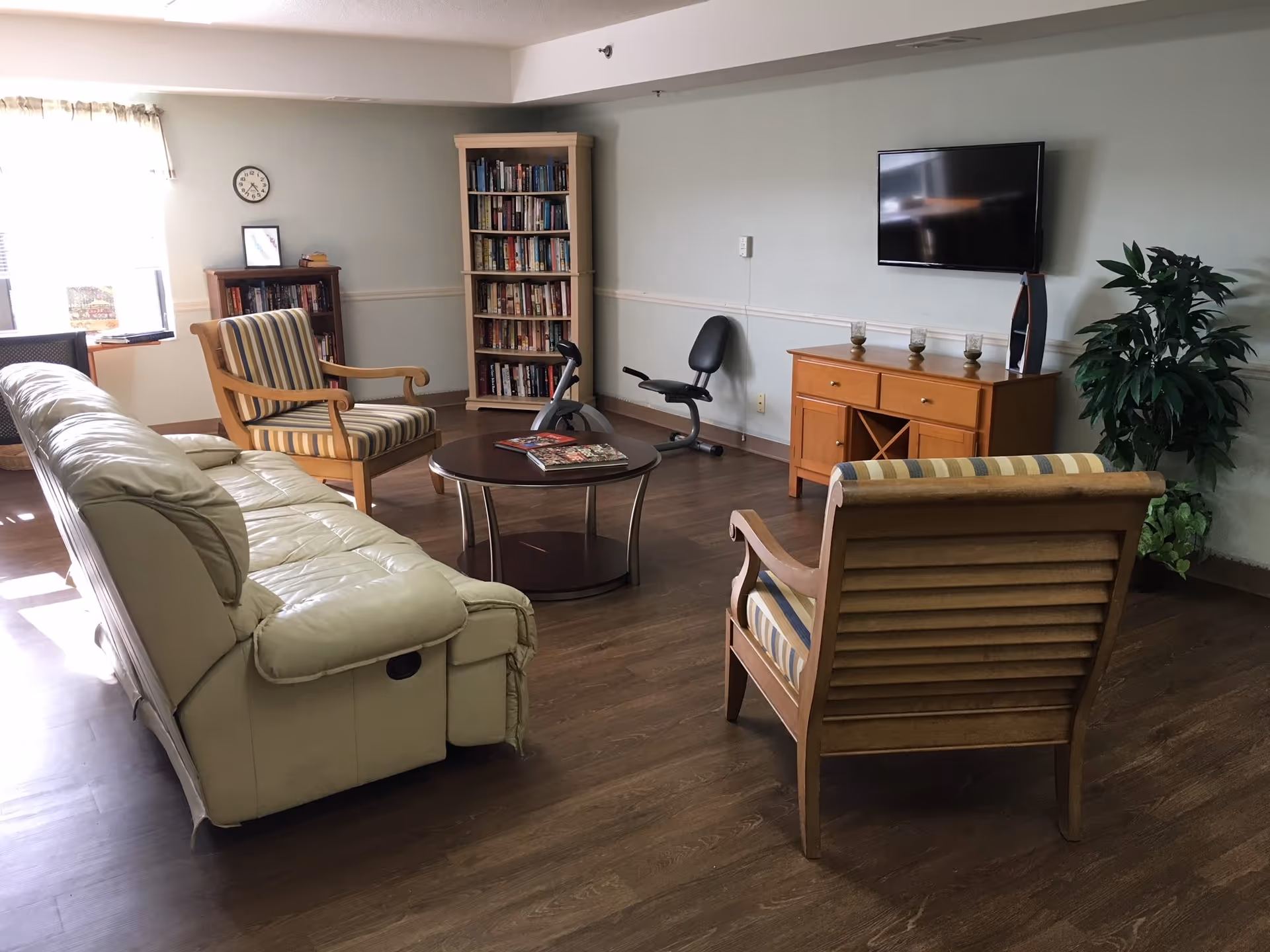A cozy living room with a cream leather sofa, two wooden armchairs with striped cushions, a round coffee table with books on it, a wooden cabinet with three candles and a decorative item, a flat-screen TV mounted on the wall, a small exercise bike, two bookshelves filled with books, a clock on the wall, and a potted plant in the corner. Sunlight is coming through a window with sheer curtains.