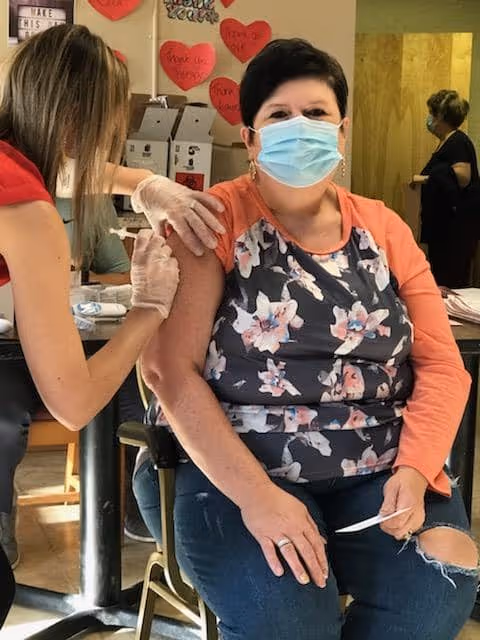 A woman wearing a floral patterned shirt and a face mask is seated while receiving a vaccination shot in her upper arm from a healthcare worker wearing gloves. The setting appears to be an indoor facility with heart-shaped decorations on the wall in the background.