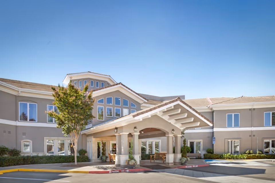 Exterior view of a two-story senior living facility building with a covered entrance, large windows, and a tree in front under a clear blue sky.