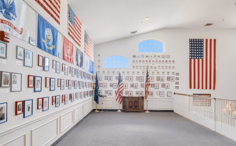 Interior hallway or gallery space with walls covered in framed photographs and military flags, including multiple American flags. Two large arched windows let in natural light, and there is a wooden podium with two American flags on stands in front of the far wall. The space has white walls, a gray carpeted floor, and a white railing on the right side.