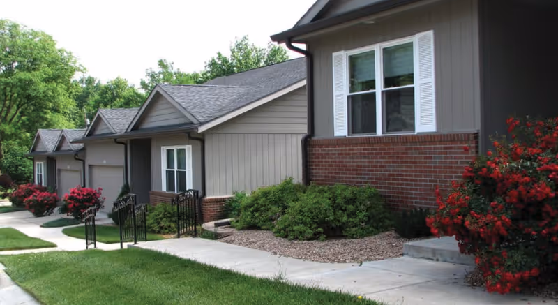 Exterior view of single-story townhouse-style units with garages, walkways, and landscaped shrubs and red flowering bushes.