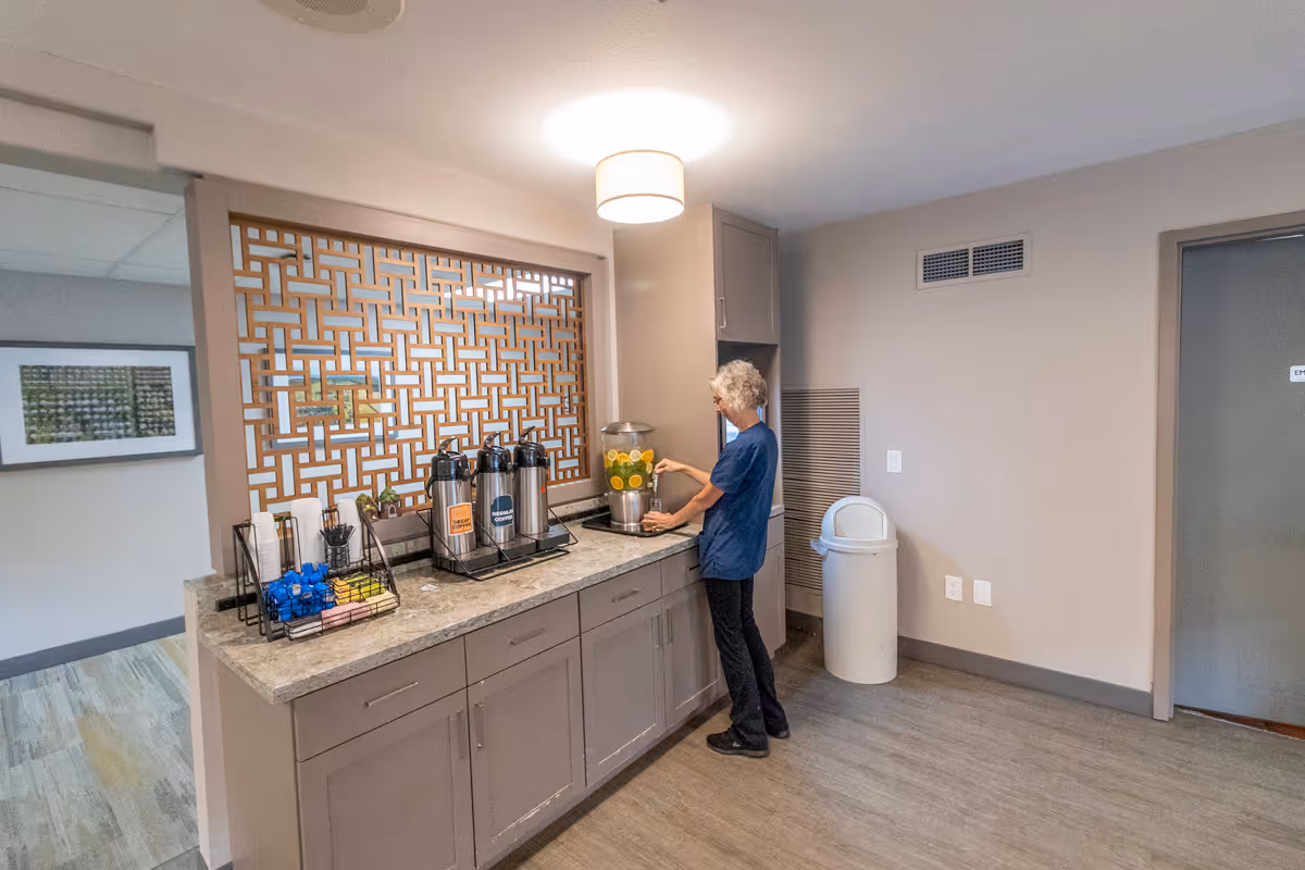 A person standing in a refreshment area with a countertop holding beverage dispensers labeled iced tea and regular coffee, along with a container of water infused with lemon slices. The area has gray cabinets, a decorative wooden lattice window, and a trash can nearby.