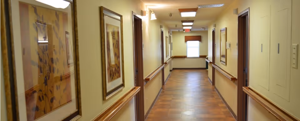 A well-lit hallway in a senior living facility with framed artwork on the walls, wooden handrails on both sides, and several closed doors leading to rooms. The floor has a wood-like finish and there is a window at the end of the corridor letting in natural light.