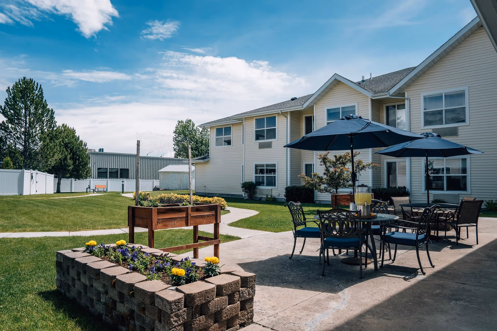 Outdoor patio area at Kalispell Assisted Living featuring a paved seating area with metal tables and chairs under large blue umbrellas, surrounded by green grass, flower beds, and a two-story beige building in the background under a partly cloudy sky.