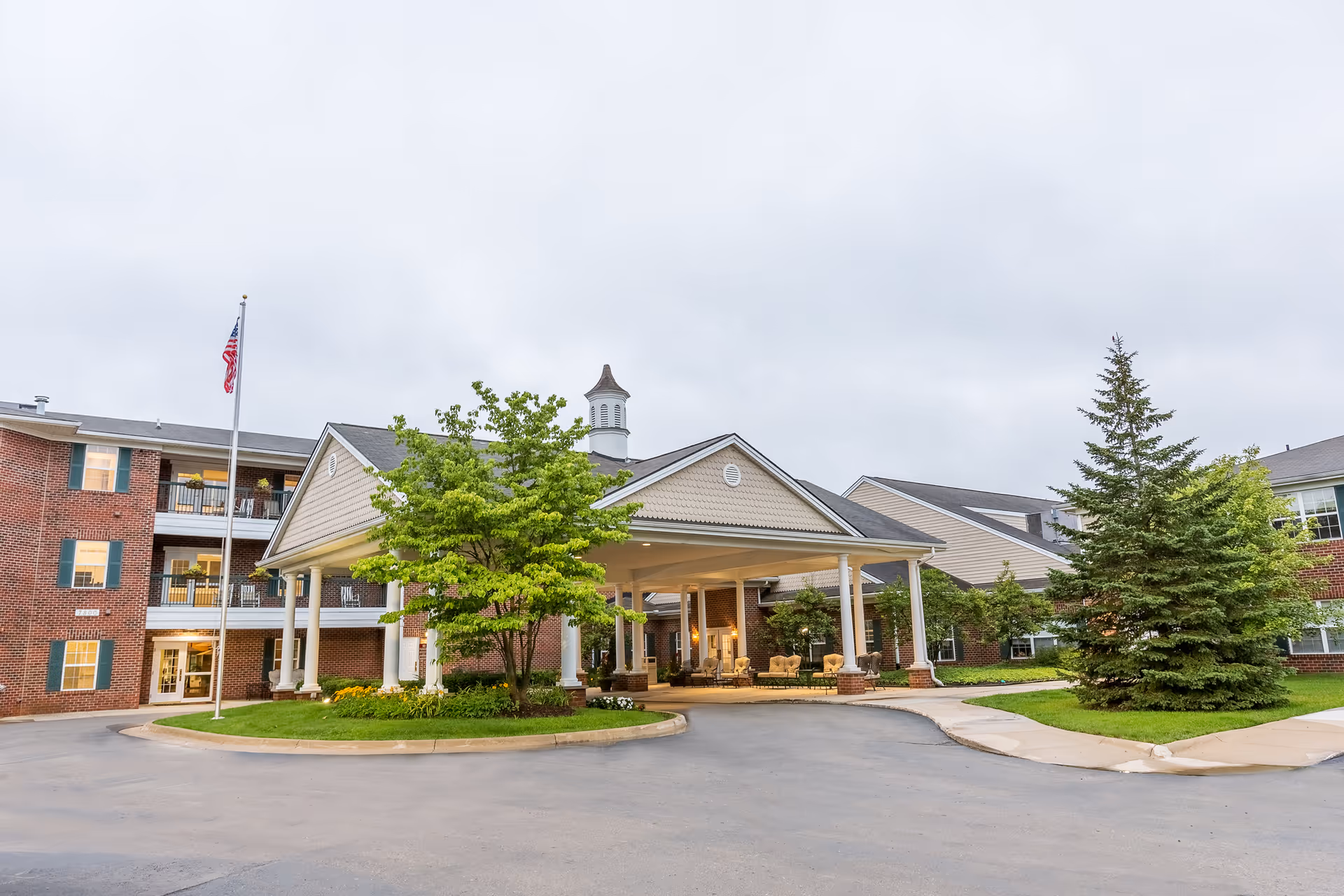 Front entrance and porte-cochere of a brick senior living building with a flagpole, trees, and outdoor seating.