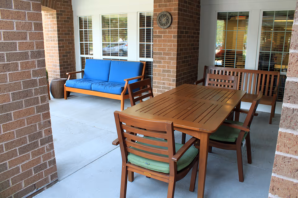 Covered outdoor patio with a wooden dining table and chairs and a blue-cushioned bench by brick columns and windows.