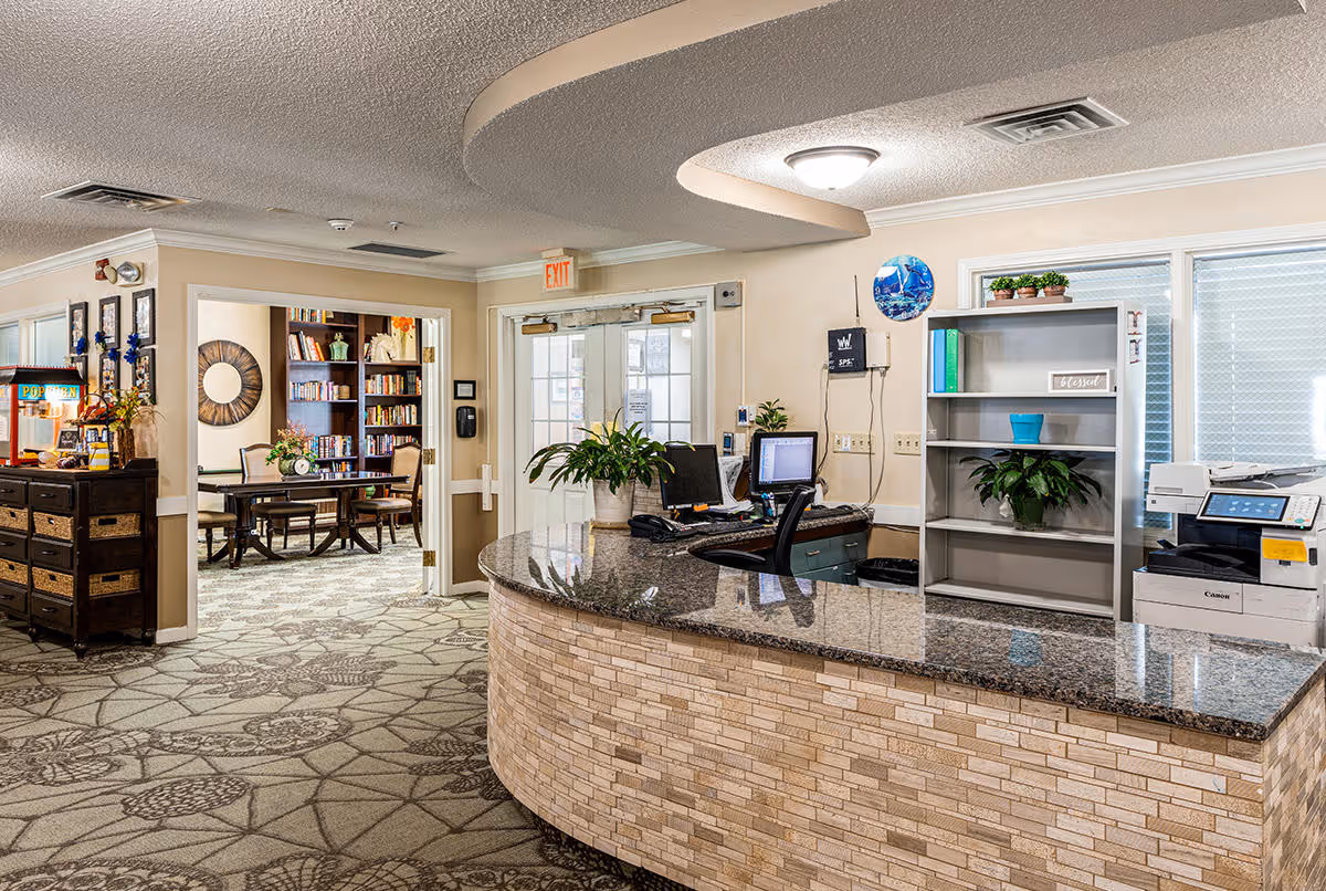 Reception area of Morton Memory Care facility featuring a curved granite countertop desk with computer monitors, a printer, and potted plants. Behind the desk is a shelving unit with decorative items and a window with blinds. To the left, there is a hallway leading to a room with bookshelves, a round mirror, and a table with chairs. The floor is carpeted with a patterned design, and the ceiling has a curved architectural feature with recessed lighting.