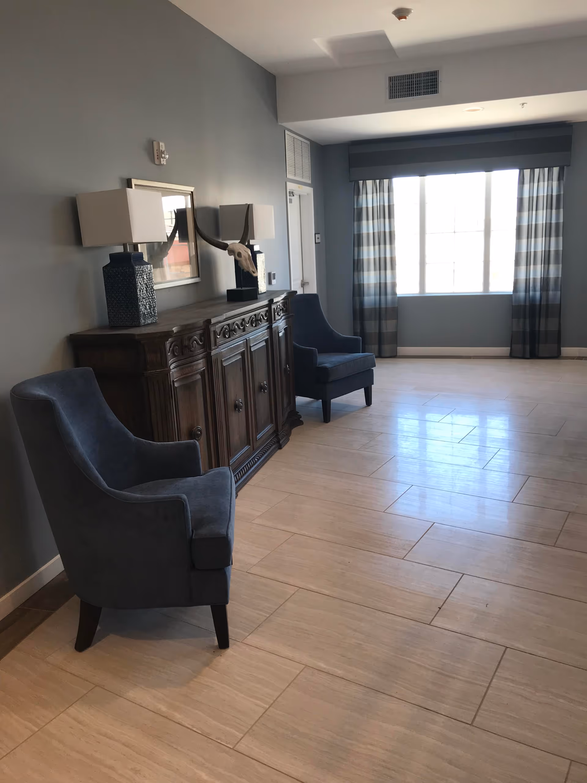 A spacious room with light-colored tiled flooring and gray walls. Two blue upholstered armchairs are positioned near a dark wooden sideboard with decorative carvings. On top of the sideboard are two table lamps with white shades and a decorative skull with horns. A large window with striped curtains allows natural light to fill the room.