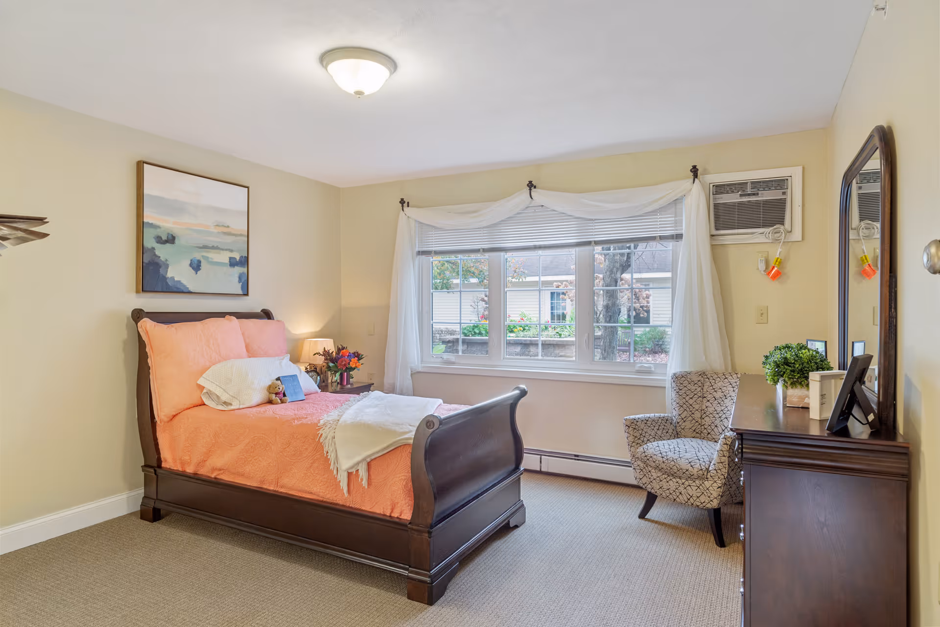 A cozy bedroom with a wooden bed frame featuring coral bedding and pillows, a small teddy bear on the bed, a nightstand with a lamp and flowers, a patterned armchair near a large window with white curtains, and a wooden dresser with a mirror and decorative items.