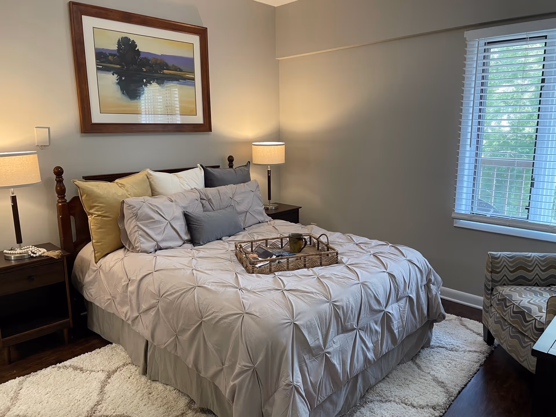A neatly made bed with a beige comforter and multiple pillows in a bedroom. There are two bedside tables with lamps on each side of the bed. A framed landscape painting hangs above the bed. A window with white blinds is on the right wall, and a patterned armchair is partially visible near the window. A light-colored rug is on the dark wooden floor.