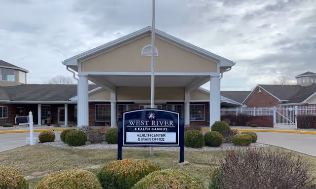 Front exterior view of West River Health Campus building with a covered entrance supported by white columns, a sign in front reading 'West River Health Campus Health Center & Main Office', surrounded by trimmed bushes and a cloudy sky overhead.