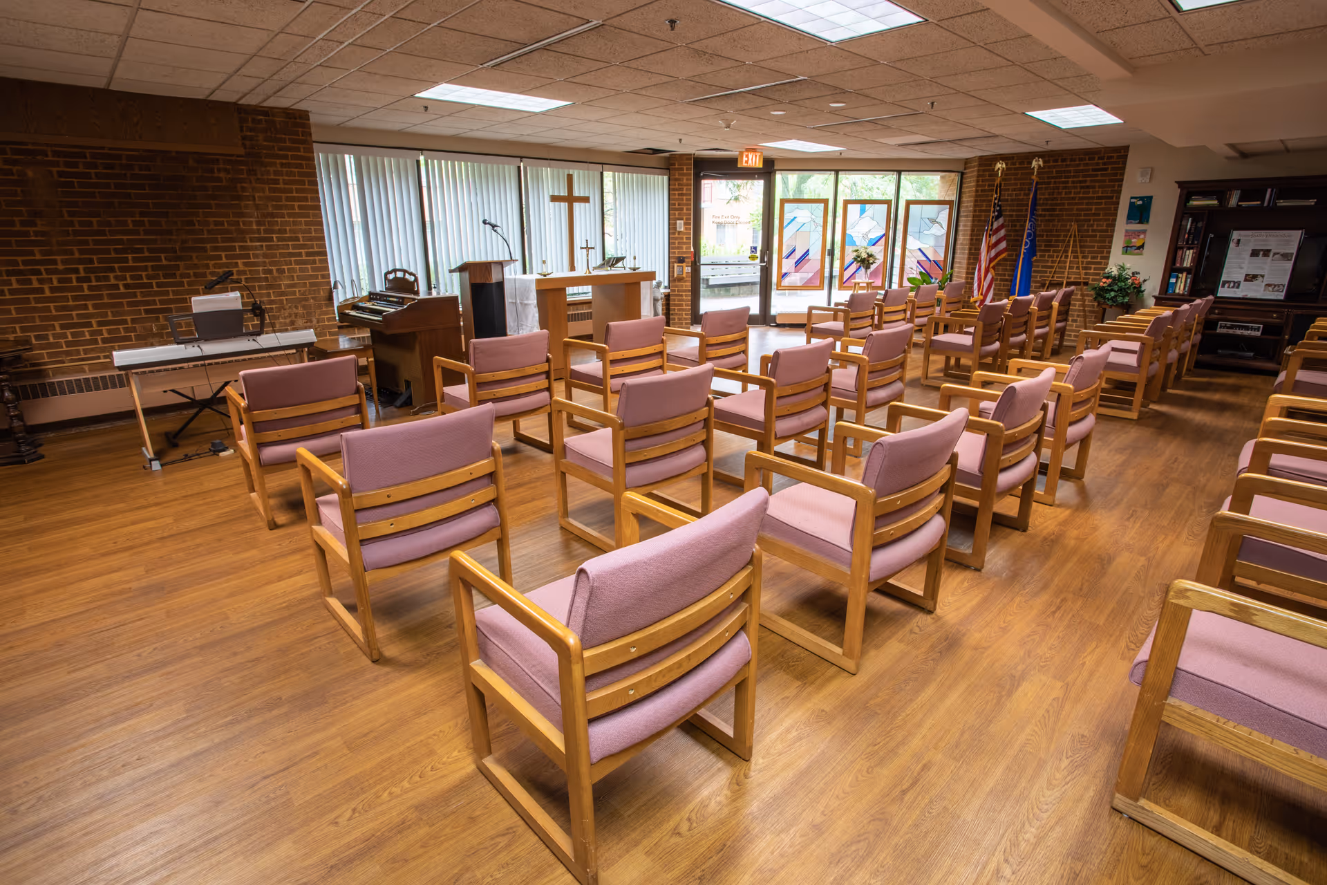 A chapel-style meeting room with rows of wooden chairs with pink cushions facing an altar, keyboard, and stained glass doors.