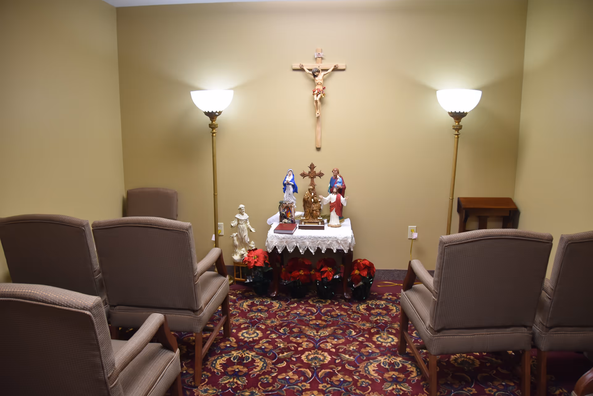 A small prayer room with six upholstered chairs arranged in two rows facing a small table covered with a white lace cloth. The table holds several religious statues and a crucifix is mounted on the beige wall above it. Two tall floor lamps with white shades flank the table, and a patterned red carpet covers the floor.