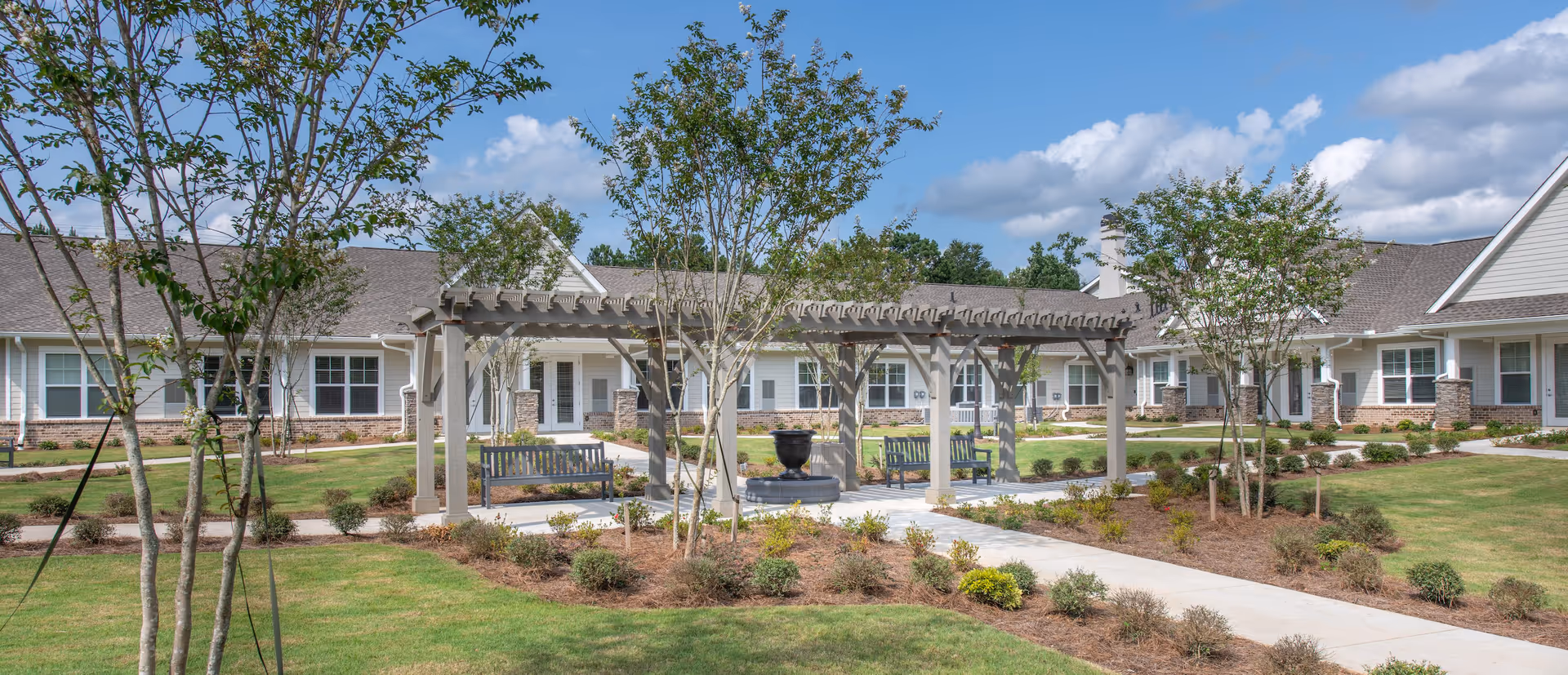 Outdoor courtyard with a pergola, benches, landscaped paths, and a single-story senior living building under a blue sky.