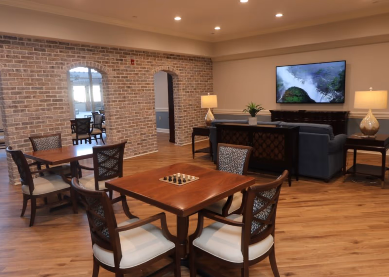 Spacious common lounge with wooden game tables and chairs, a sofa seating area with a wall-mounted TV, and an exposed brick wall with arched openings.