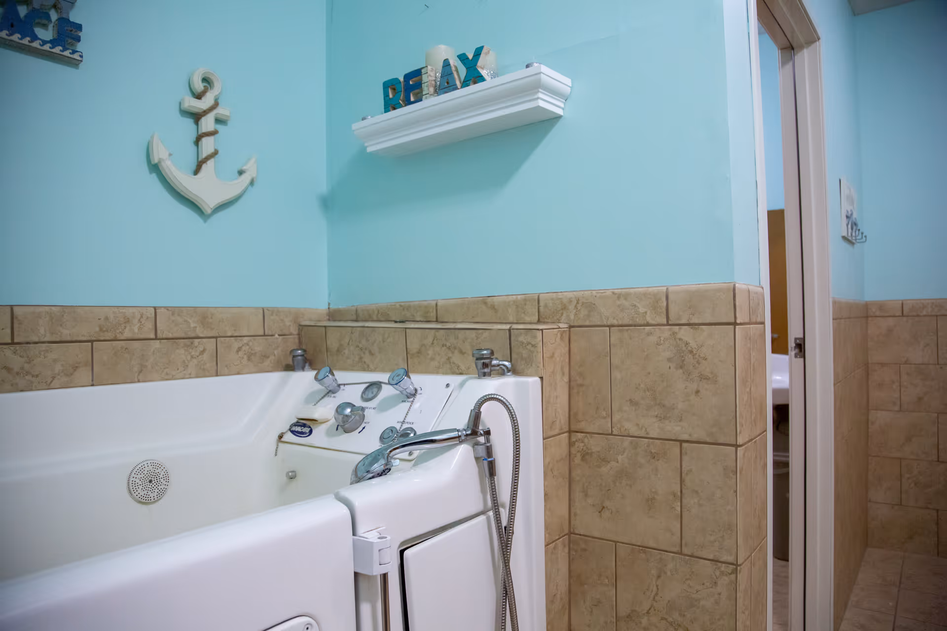 A walk-in bathtub with control knobs and a handheld showerhead in a bathroom with light blue walls and beige tiled wainscoting. Wall decorations include a white anchor and a shelf with the word RELAX and candles.
