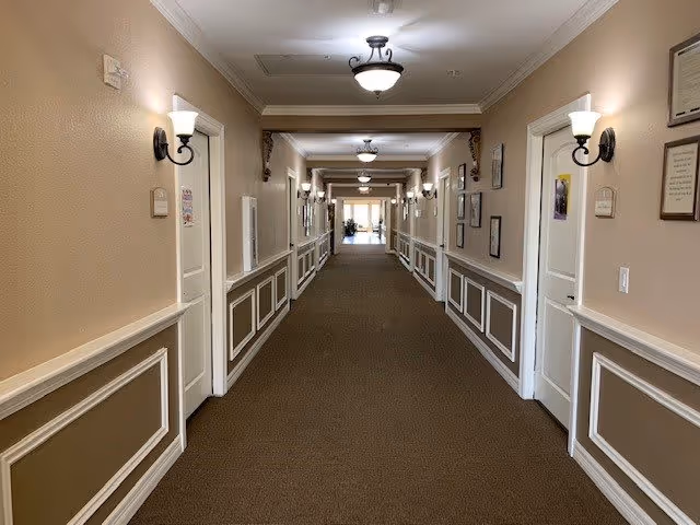 Long indoor hallway in a senior living facility with beige walls, white trim, multiple closed doors on both sides, wall-mounted light fixtures, ceiling lights, and framed pictures on the walls. The hallway is carpeted and leads to a brighter area at the far end.
