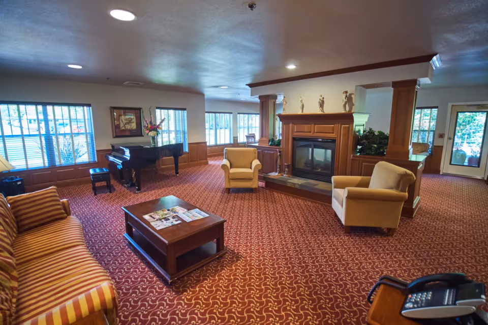 A spacious living room area with patterned red carpet, a striped sofa, two beige armchairs, a wooden coffee table with magazines, a grand piano near windows with blinds, and a fireplace with decorative figurines on the mantel. There is a door leading outside and some plants near the fireplace.