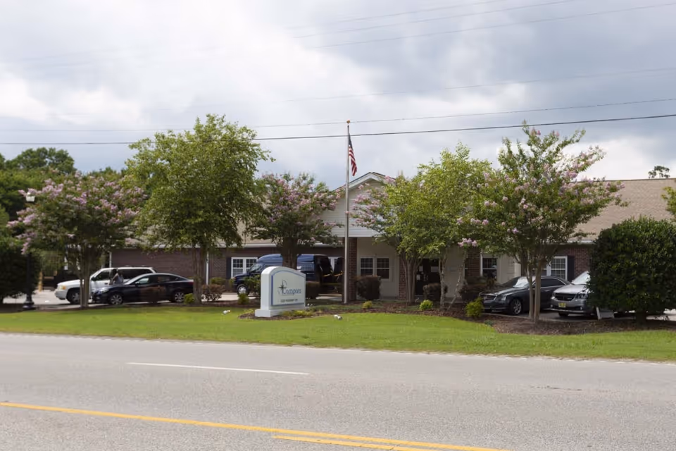 Front exterior of the Compass Post Acute Rehabilitation building with a sign, flagpole, parked cars and trees along the road.