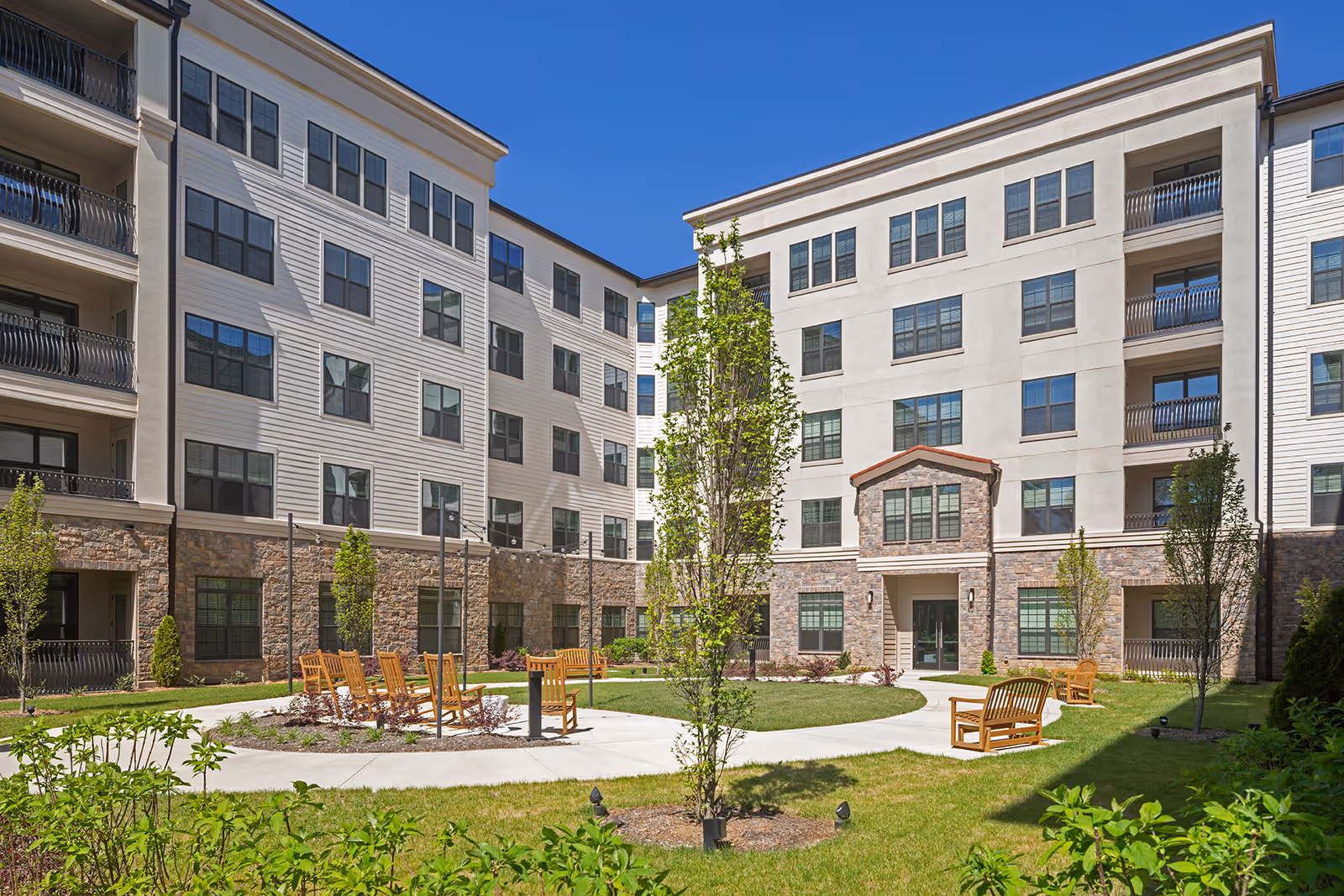 Outdoor courtyard area of a senior living facility with a circular paved walkway, wooden benches, young trees, and a multi-story building with balconies and large windows surrounding the courtyard under a clear blue sky.