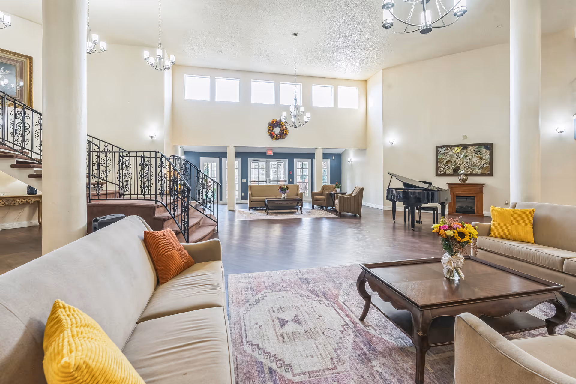 A spacious and well-lit living room area with high ceilings, large windows near the ceiling, and multiple seating arrangements including beige sofas with yellow and orange pillows. There is a wooden coffee table with a vase of flowers, a grand piano near a fireplace, and a staircase with ornate black railings leading to an upper floor.