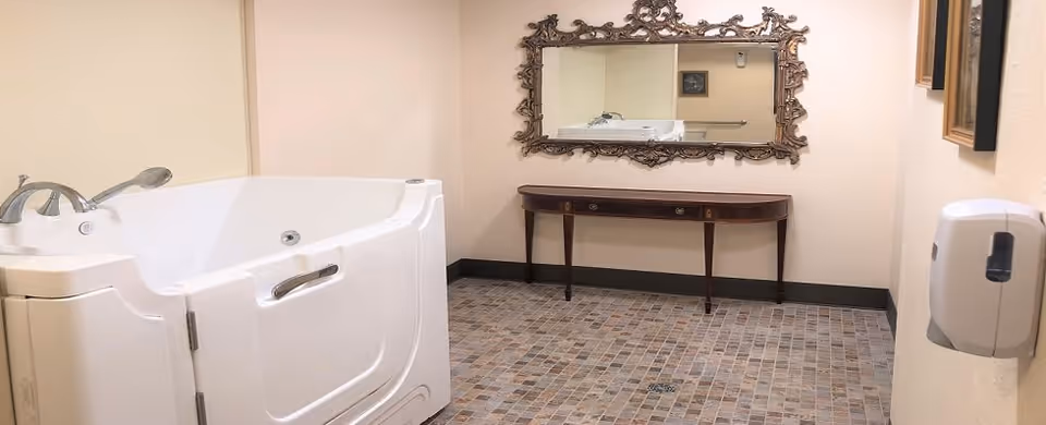 A spacious bathroom featuring a large white walk-in bathtub with a door and grab bar on the left side. The floor is covered with small square tiles in various shades of brown and beige. On the far wall, there is an ornate, decorative mirror above a narrow wooden console table with three drawers. A soap or sanitizer dispenser is mounted on the right wall.