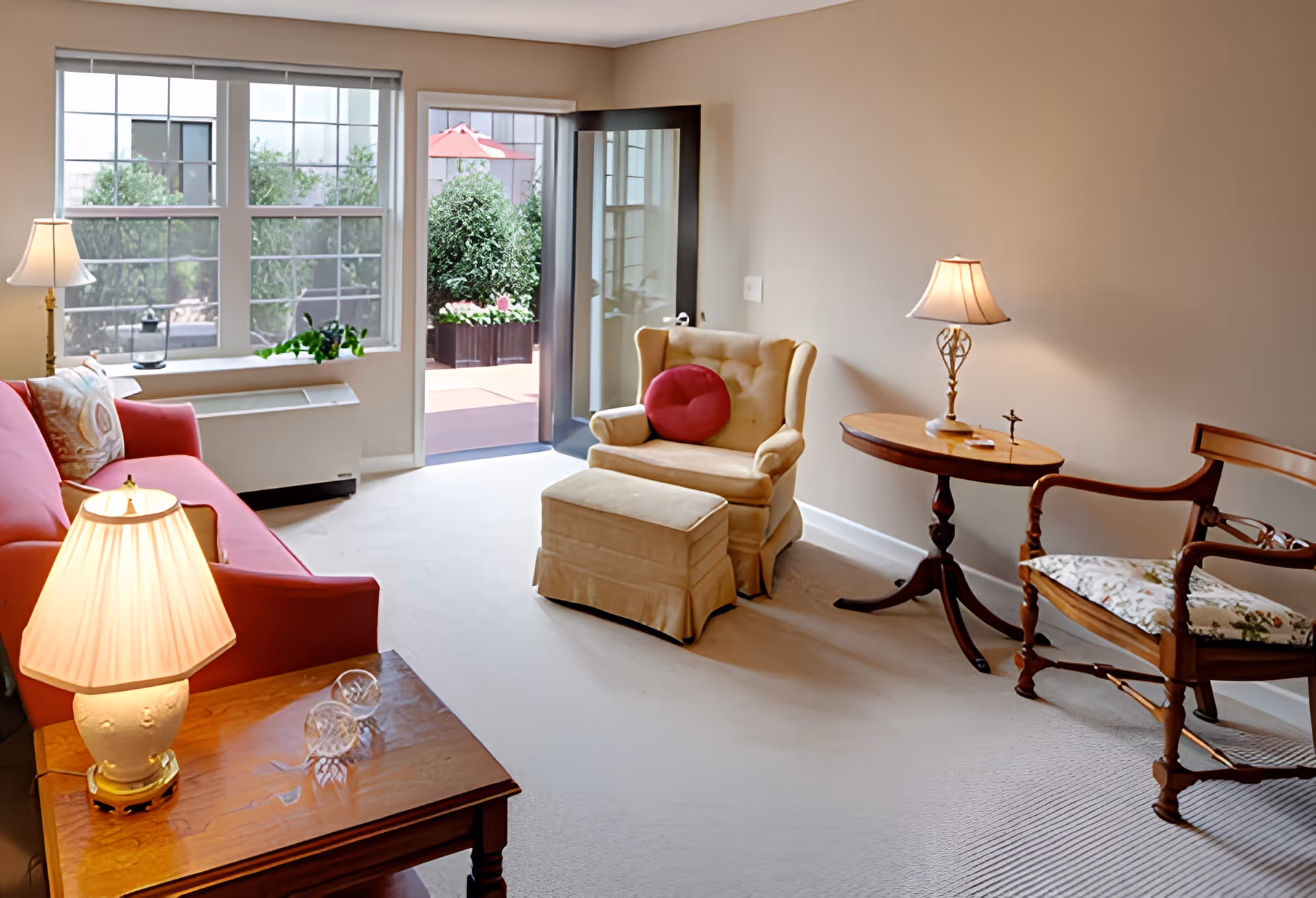 A cozy living room with a red sofa, a beige armchair with a red round pillow, a wooden side table with a lamp, and a wooden chair with floral upholstery. There is a glass door open to an outdoor patio with greenery and a red umbrella visible through the windows.