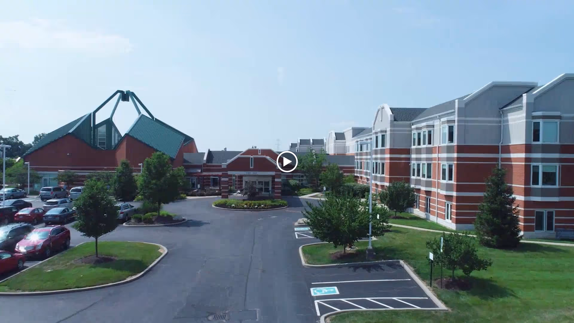 Exterior view of a senior living facility with a large parking lot, landscaped greenery, and a multi-story building with red brick and white trim under a clear blue sky.