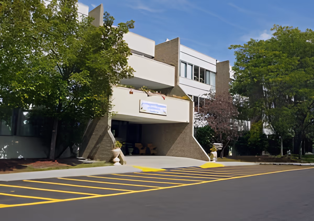 Front entrance of Angel Gardens Assisted Living showing a covered drop-off, landscaped trees, and a yellow-striped roadway.