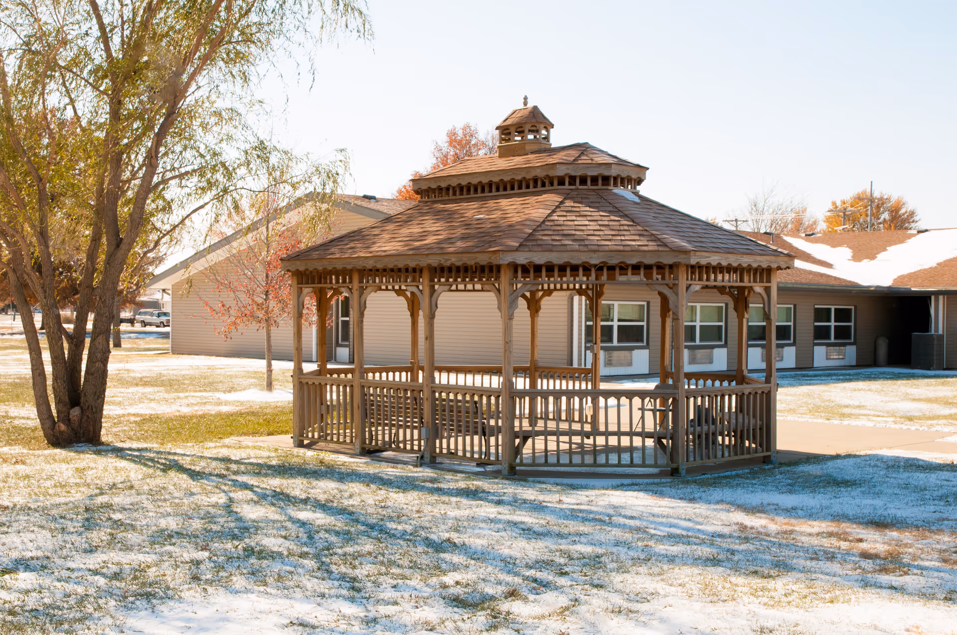 Wooden gazebo on a lightly snow-covered lawn in front of a single-story senior living building.