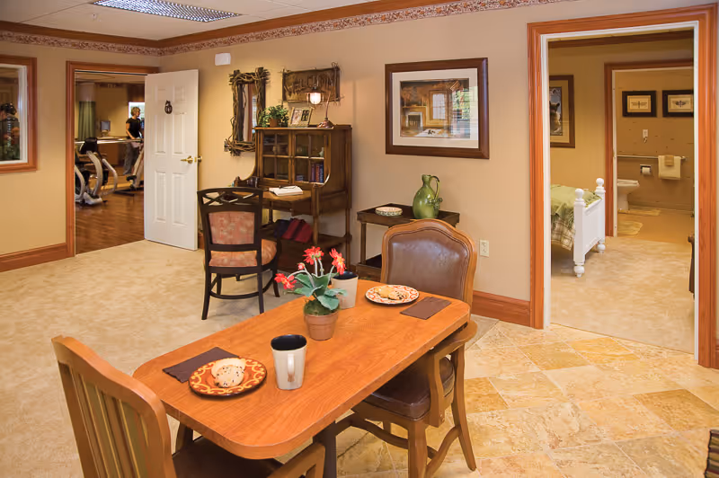 A cozy interior room with a wooden table set for two, featuring plates, a mug, and a small potted plant with red flowers. Behind the table, there is a wooden desk with a chair and decorative items on the wall. Two doorways are visible: one leads to a bedroom with a white bed frame, and the other opens to a room with exercise equipment and a person walking on a treadmill. The room has beige walls, tiled and carpeted floors, and framed artwork on the walls.
