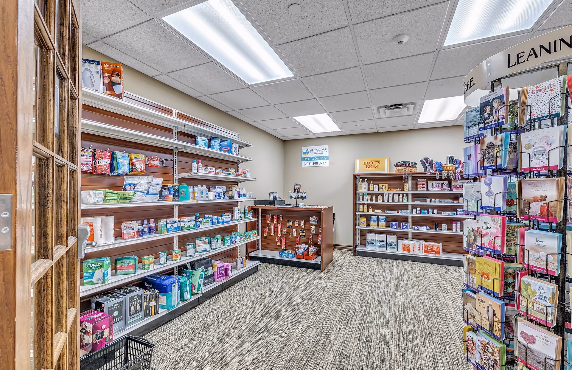 Interior view of a small retail shop area within a facility, featuring shelves stocked with various health and personal care products, greeting cards on a rotating display rack, and a counter with additional items. The room has a carpeted floor, fluorescent ceiling lights, and a wooden door partially open on the left.