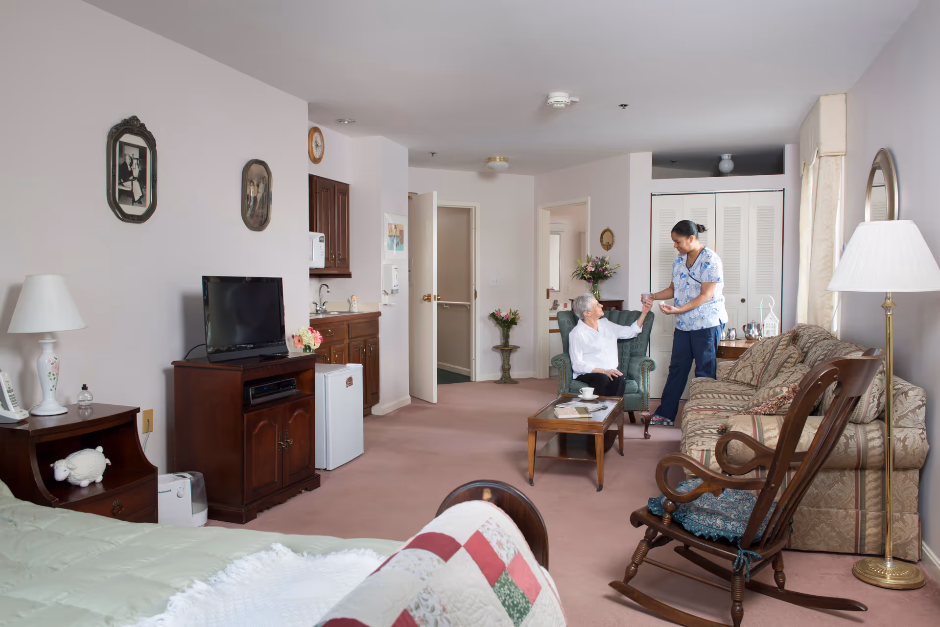 A senior woman sitting in a green armchair in a cozy living area of a senior living facility, receiving a glass of water from a caregiver. The room features a bed with a quilt, a wooden rocking chair, a sofa, a coffee table, a TV on a wooden stand, a small kitchenette area, and soft pink carpeting.