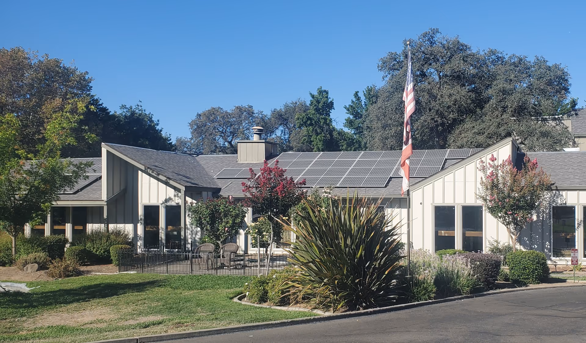 Single-story light-colored building with solar panels on the roof, an American flag, outdoor seating, and landscaped grounds.