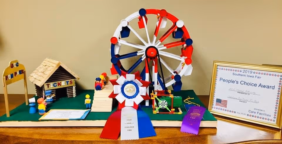 A miniature model of a fun park featuring a Ferris wheel, a ticket booth, and small figurines. The model is decorated with red, white, and blue colors and includes award ribbons and a framed certificate for the 2019 Southern Iowa Fair People's Choice Award in the Care Facilities division.