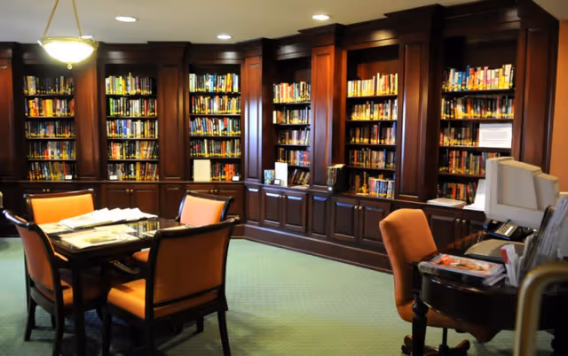 A cozy library room with dark wooden bookshelves filled with books lining the walls. In the center, there is a table with four orange cushioned chairs around it. To the right, there is a desk with an orange chair and a computer monitor. The room has a green carpet and warm lighting from ceiling fixtures.