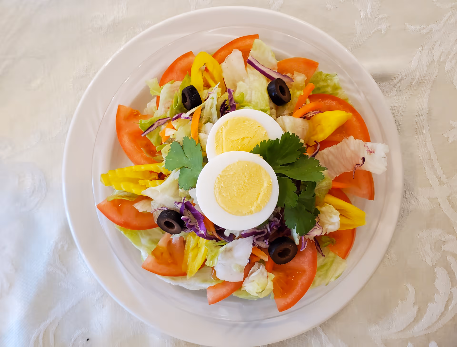 A fresh salad on a white plate featuring sliced tomatoes, yellow bell peppers, black olives, shredded purple cabbage, lettuce, and two halves of a hard-boiled egg garnished with cilantro, placed on a white textured tablecloth.