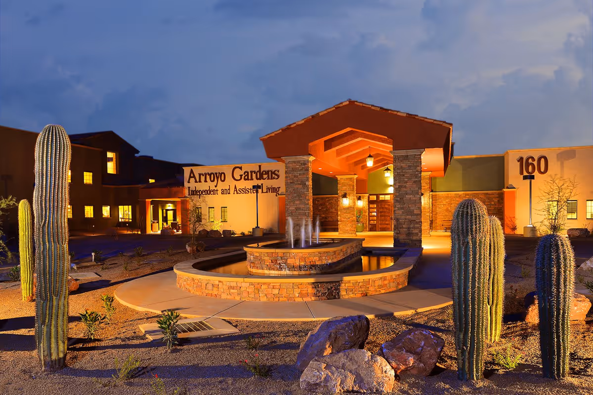 Entrance of Arroyo Gardens senior living building at dusk with a fountain, illuminated porte-cochere, and desert landscaping including tall cacti.