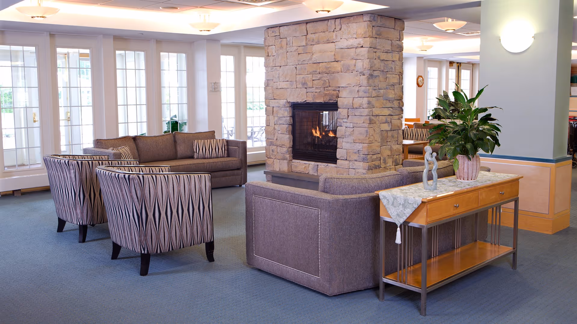 A cozy living room area in a senior living facility featuring a stone fireplace with a fire burning inside. The room has large windows letting in natural light, a gray sofa, two patterned armchairs, and a wooden console table with a potted plant and decorative sculpture on top.