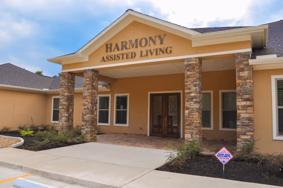 Front exterior view of Harmony Assisted Living facility with a covered entrance supported by stone pillars, beige walls, and a paved walkway leading to double wooden doors.
