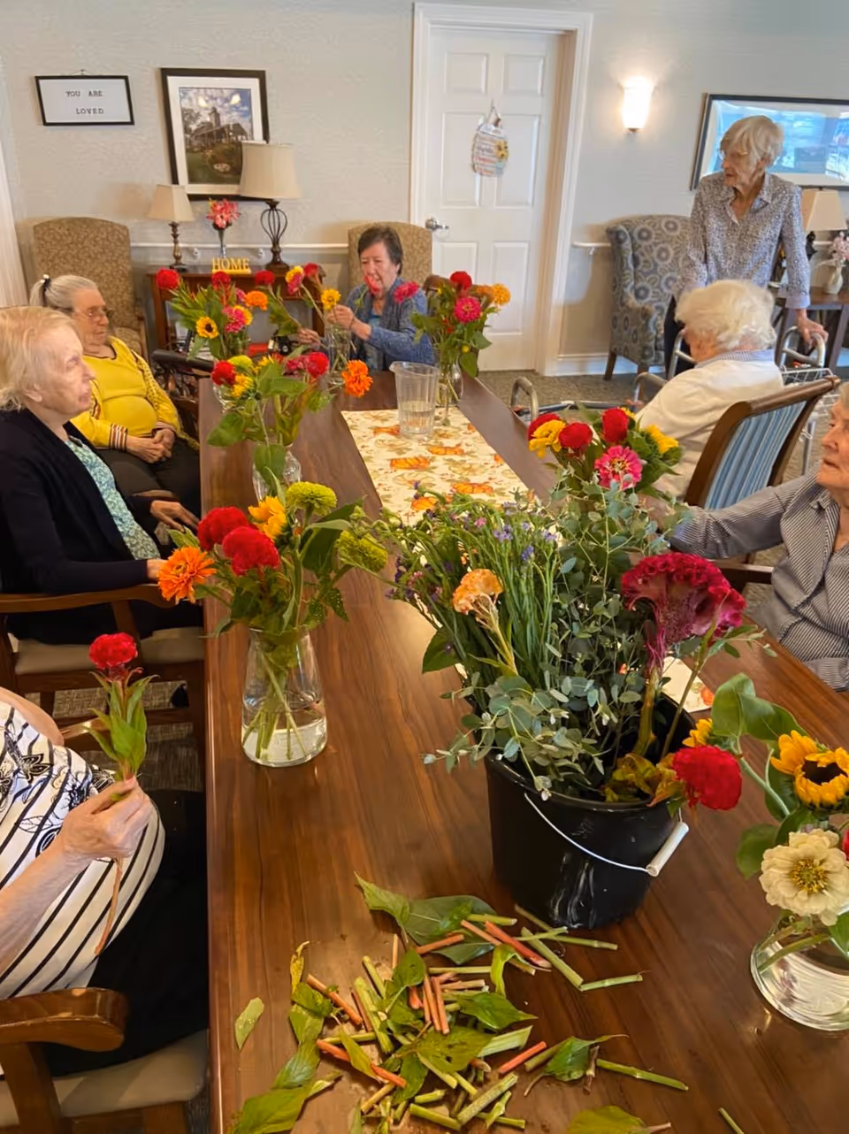 A group of elderly women sitting around a long wooden table in a well-lit room, arranging colorful flowers in vases. The table has a floral runner and flower stems and leaves scattered on it. The room has comfortable chairs, framed pictures on the walls, and a door in the background.