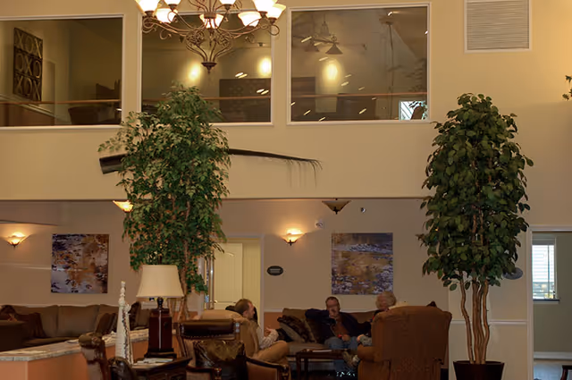A cozy living room area in an assisted living facility with several people sitting and conversing on sofas and armchairs. The room features two large potted plants, wall-mounted lights, framed artwork, and a chandelier hanging from the ceiling. There is a second-floor balcony with windows overlooking the space.