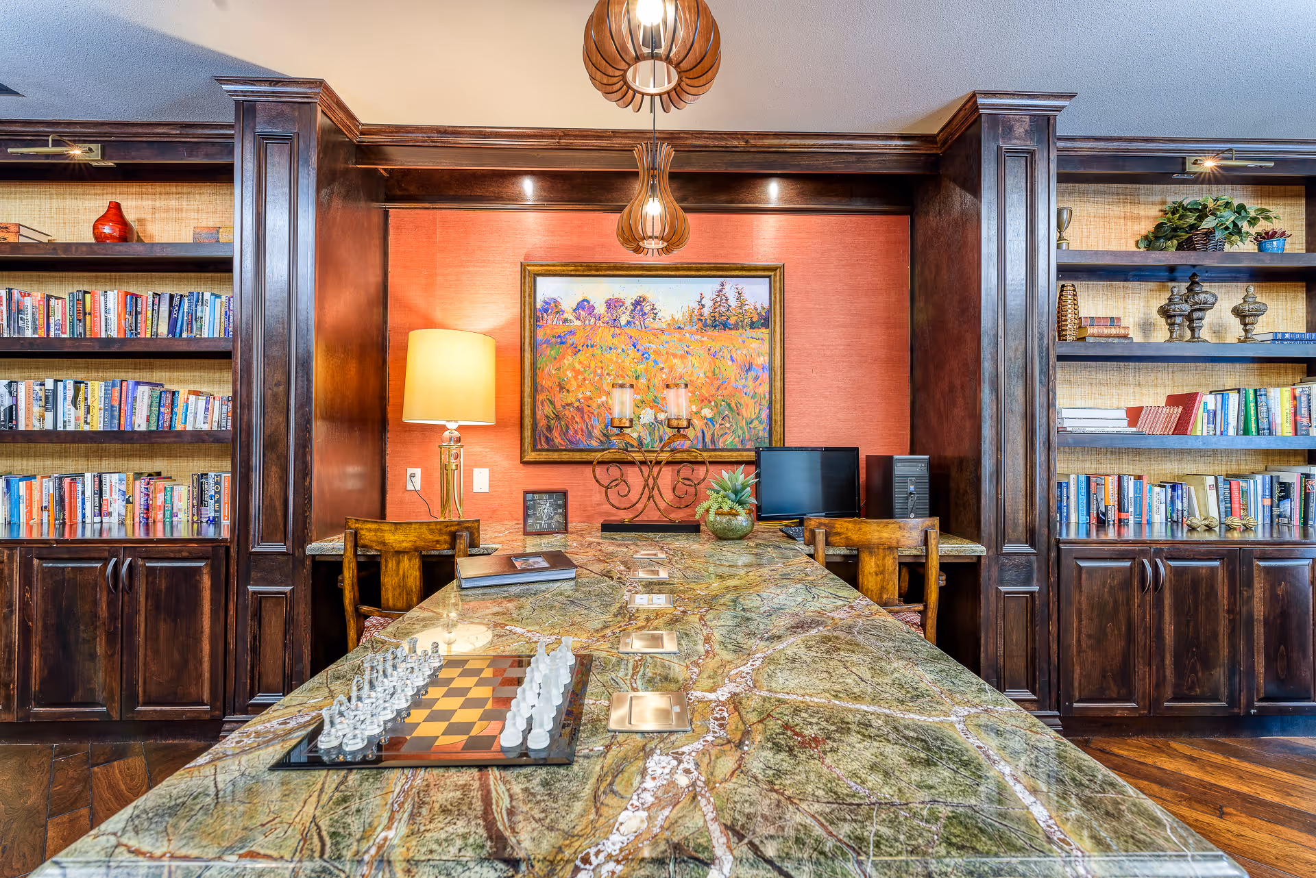 Communal library-style room with a long marble table holding a chess set, bookshelves, artwork, lamps and pendant lights.
