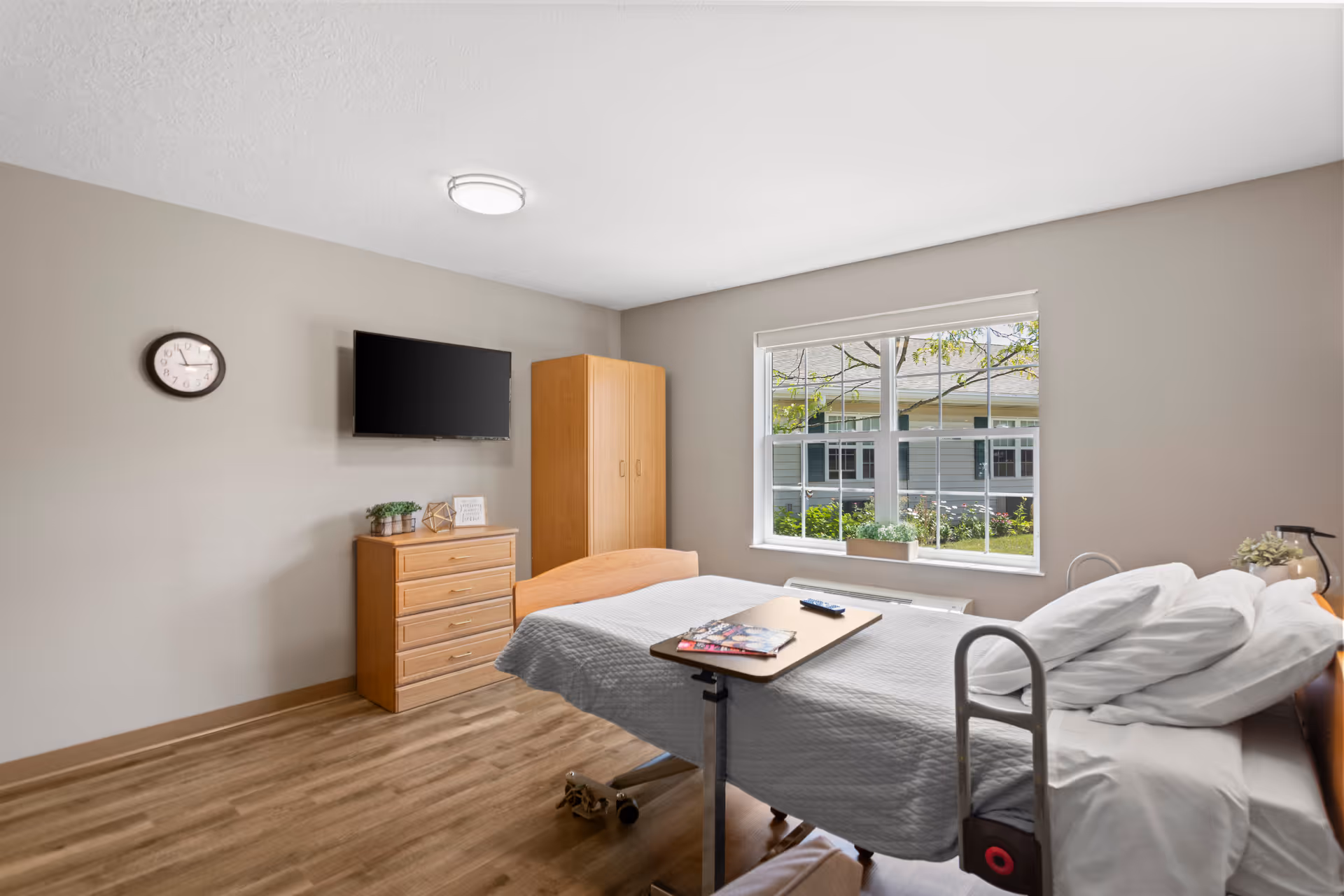 A bright and clean nursing facility bedroom with a single bed covered in a gray quilt, a wooden dresser and wardrobe, a wall-mounted flat screen TV, a clock on the wall, and a large window showing greenery outside.