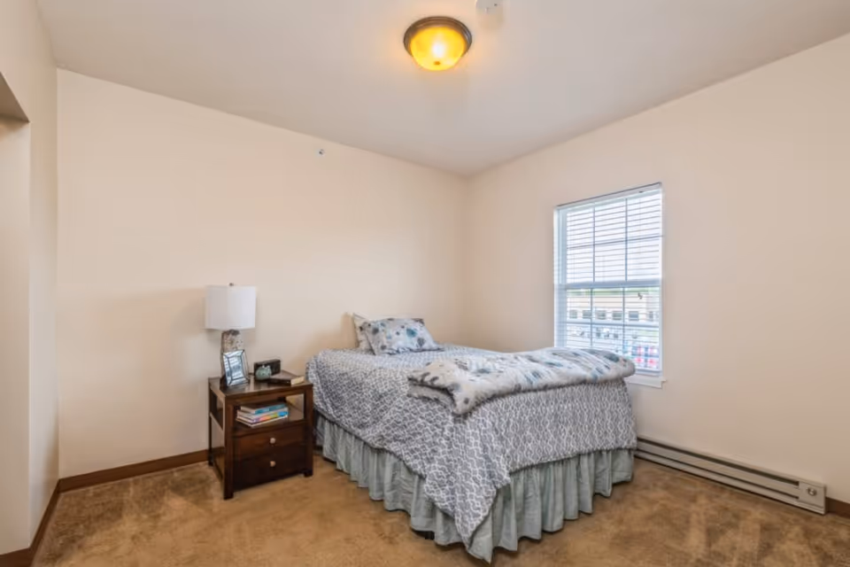 A simple bedroom with beige walls and carpet, featuring a single bed with patterned bedding and a floral pillow. Next to the bed is a wooden nightstand with a lamp, a framed photo, and some books. A window with white blinds lets in natural light.